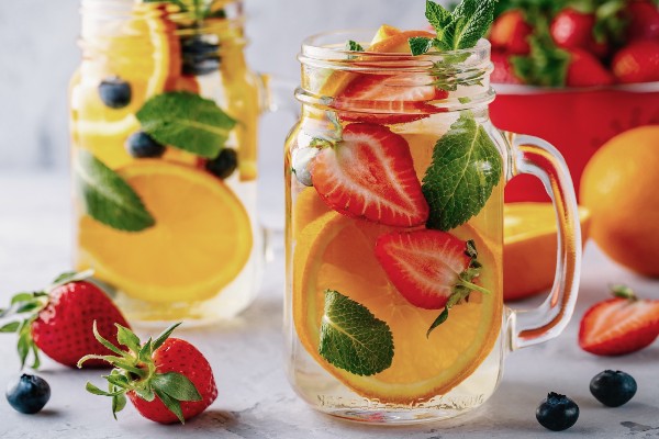 2 glasses with handles filled with water, orange slices, , mint leaves, with strawberries, oranges and blueberries in the background