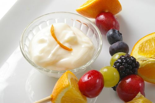 small glass bowl filled with yogurt fruit dip surrounded by an assortment of fruit on a stick
