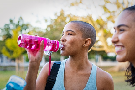 dark skin lady in light blue tank top with short dark hair drinking out of a pink water bottle with trees in the background