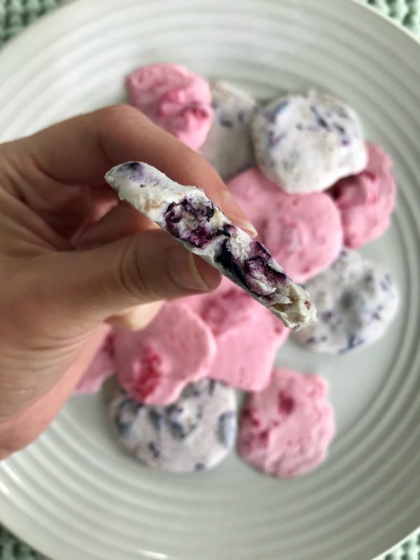 Close up of a girl's hand holding a yogurt drop that has a bite out of it with a backdrop of a plate of strawberry and blueberry yogurt drops