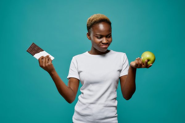 Front view of african woman in white shirt keeping tasty chocolate plate in one hand and healthy apple in another on blue isolated background. Serious girl choosing between good and bad food.