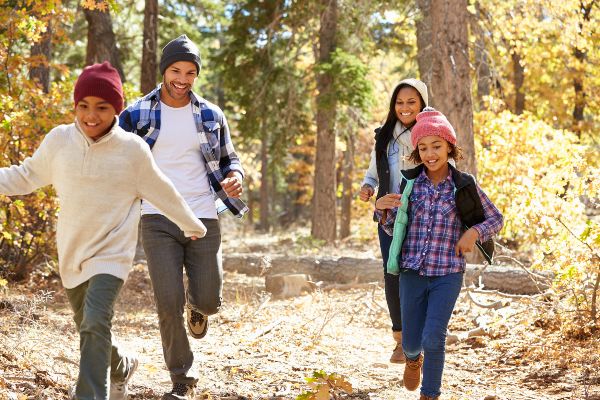 Family of two adults and two children in seasonal clothing run through a forest of autumn foliage