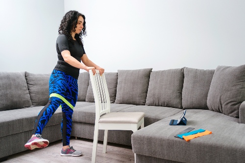 Girl with dark curly hair and blue leggings holds onto the back of a white chair in her living room and looks at a tablet screen to follow a workout