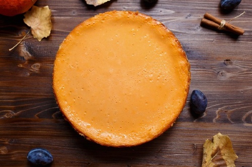 Whole orange cheesecake sits on wooden table surrounded by fruit, cinnamon sticks, and fall leaves