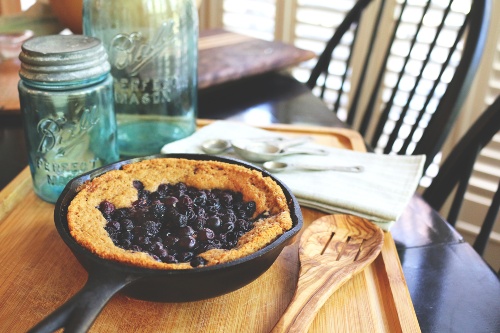 Berry cake in cast iron skillet on top of a wooden cutting board with a cloth napkin, wooden spoon, and blue mason jars