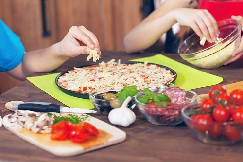 Kids hands adding cheese to a small pizza with toppings surrounding them