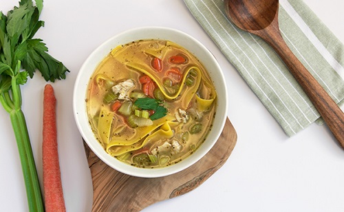 bowl of veggie filled chicken noodle soup sits on a wooden board surrounded by a napkin, spoon, and veggies.