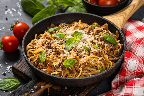 skillet of spaghetti with meat, cheese, and basil surrounded by fresh veggies and a checkered tablecloth