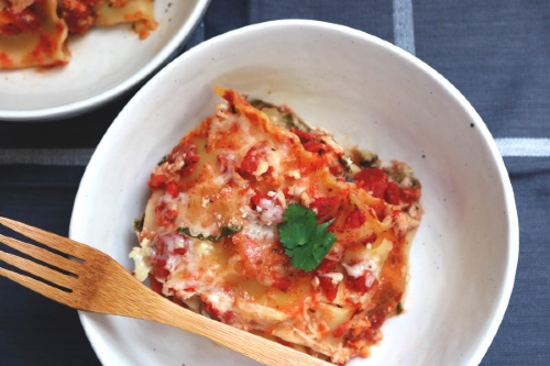Square of lasagna sits in a white bowl with a wooden fork resting on the side