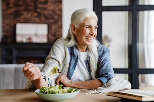 Smiling senior woman reading a book while sitting in the kitchen, eating salad