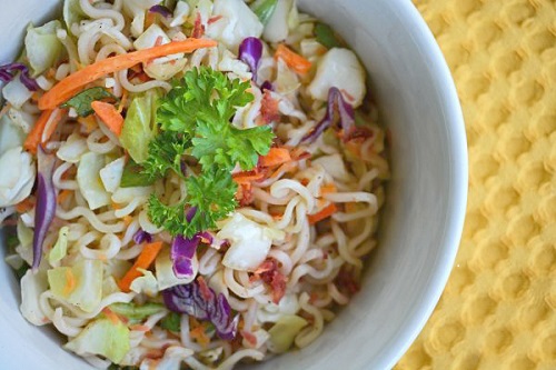 Colorful bowl of coleslaw sits on top of ramen noodles against a yellow background