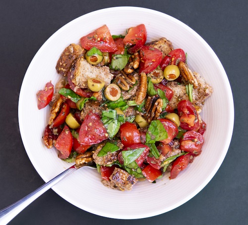 colorful salad with bread, tomatoes, olives, greens, and pecans in a white bowl with a silver spoon