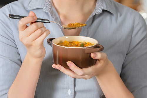 Woman in blue shirt holds up brown bowl of lentil soup and a spoonful of lentils