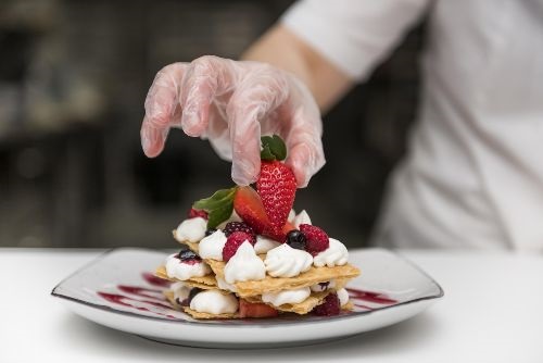 chef hand with glove putting strawberries on top of a stack of graham crackers sandwiched with whipped cream on a white plate