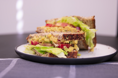 Sandwich with chickpea salad, lettuce, and tomatoes on two slices of whole wheat bread, on a purple napkin.