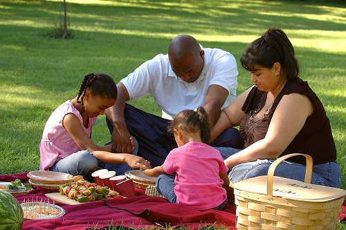 Family of four sit on a picnic blanket with food and hold hands to give thanks.