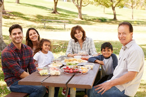 Family of four adults and two kids sit at a picnic table and smile with containers of food on the table