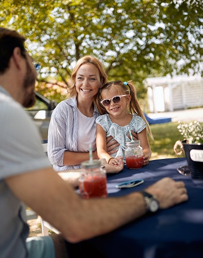 Mom and daughter sit at a shaded picnic table with bright drinks. The girl is wearing sunglasses and they are smiling at a male person..