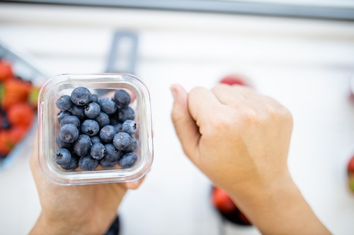 Female fist next to small plastic container full of blueberries above blurry fruit. Fresh and blurry berries in plastic containers from above.