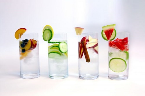 Four glasses of water in a row filled with various fruits and cucumber slices.