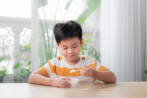 Young boy sits at a table and smiles as he scoops up yogurt from a container. He is wearing an orange striped shirt.