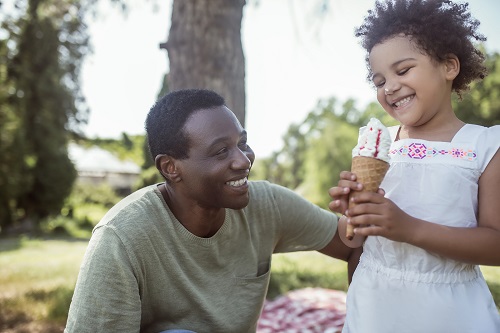 A dad kneels next to his daughter as she smiles and holds an ice cream cone