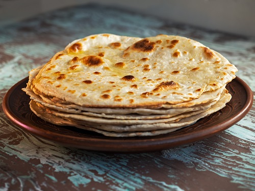 Taco tortilla cakes lay in a stack on a brown plate on a variegated wooden background