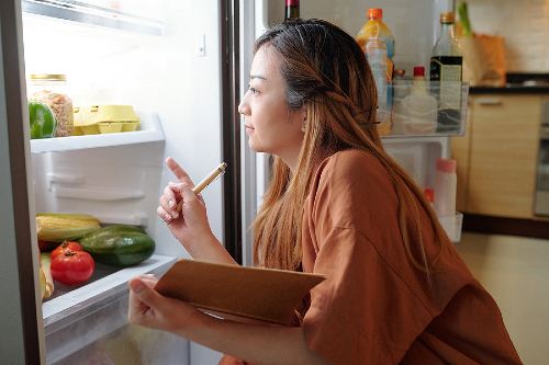 woman with a notepad looking in the refrigerator to see what food she already has