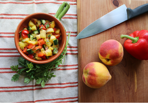 cut peaches, red pepper and green onions in a ceramic bowl sitting on top of a red and shite striped dish towel beside 2 peaches and a red bell pepper and knife on a wooden block