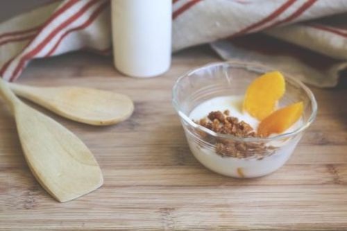 vanilla yogurt with 2 peaches and crumbled graham cracker in a glass dish on a wooden table with wooden spoons, milk and a red and white striped towel in the background