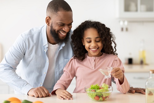 Father and daughter smile as they pour olive oil over a salad in the kitchen