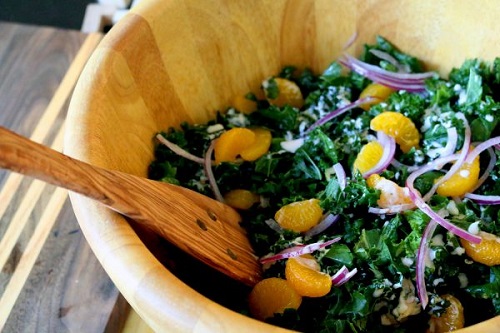 kale, red onion, and orange salad sit in a wooden bowl with a wooden serving spoon