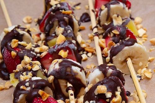 frozen strawberries, bananas and pineapple covered in chocolate and peanuts on wooden sticks
