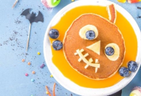 white plate with a pancake with bananas and blueberries for eyes and apples for the nose and mouth on a blue background with a black candy bat beside the plate