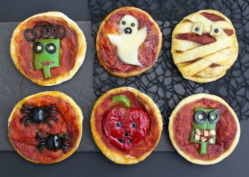 6 round pieces of dough covered with red sauce and decorated with halloween designs made of vegetables, cheese and olives on a black background