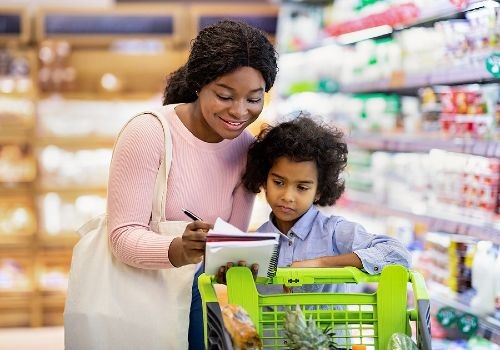 African american mom and daughter looing at a grocery list standing behind a grocery card in the middle of a grocery aisle