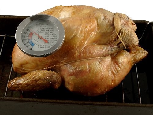 A roasted chicken with a meat thermometer stuck in the breast on a roasting pan on a wire oven rack