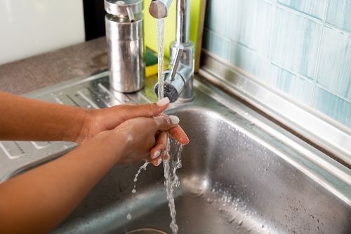 African American hands with soap on them under running water in a silver sink with a silver soap bottle beside the sink and a window behind the sink