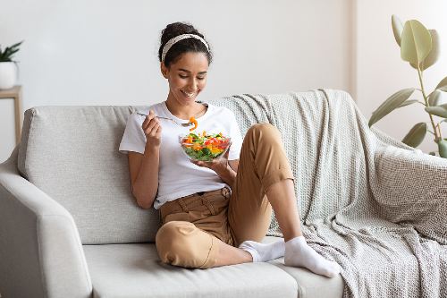 young girl smiling while eating a bowl of fresh vegetables and sitting on a couch