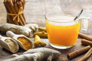 ginger tea in a glass mug with a spoon inside it and 2 scoops of spices with ginger root and a bunch of cinnamon sticks beside it