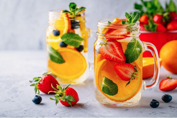2 glasses with oranges, strawberries and mint inside the glasses surrounded by fruits