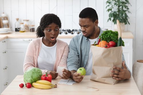 Black couple looking at a grocery receipt with expressions of shock surrounded by a bag of food and fruits and vegetables on the counter