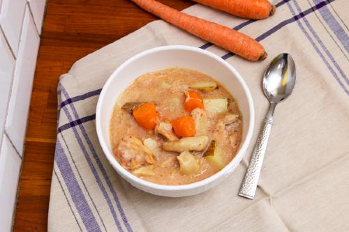 a bowl of chicken and dumplings set on a white and blue napkin with fresh carrots and a spoon resting near the bowl