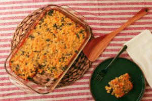 Spicy Kale Corn Pudding in a glass casserole dish with a piece of the corn pudding on a black plate beside the casserole dish