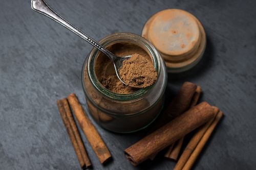 a spoon scooping ground cinnamon from a glass jar that is sitting on a dark surface with dried cinnamon sticks surrounding it 