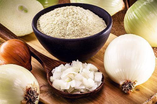 a small, black bowl of onion powder with a wooden spoon full of chopped onions sitting in front and a fresh, peeled onion sitting off to the side