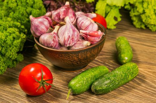 A wooden bowl filled with garlic bulbs with cucumbers , tomato and other greens beside it on a wooden surface