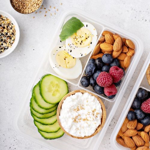 container holding almonds, berries, boiled eggs, cucumber slices and a cracker with white cream cheese spread on top