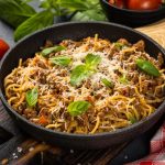 skillet of spaghetti with meat, cheese, and basil surrounded by fresh veggies and a checkered tablecloth