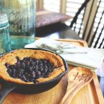 Berry cake in cast iron skillet on top of a wooden cutting board with a cloth napkin, wooden spoon, and blue mason jars